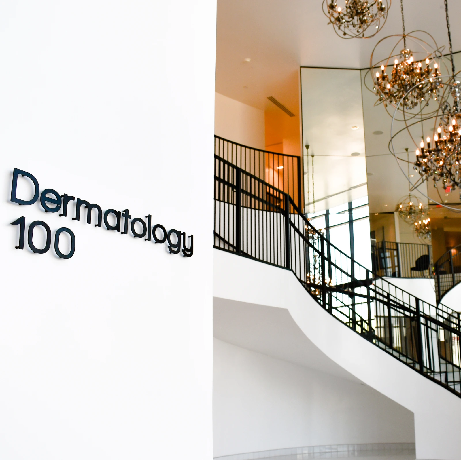 Dermatology 100 signage in a modern medical office lobby with curved staircase and gold chandeliers